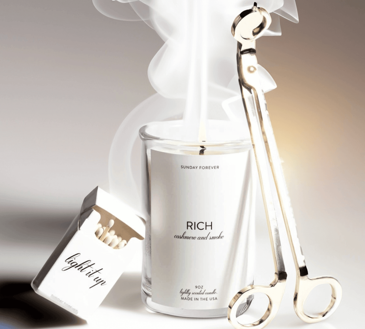 Lit Rich candle with cashmere and smoke beside a gold wick trimmer and a light it up matchbox by Sunday Forever